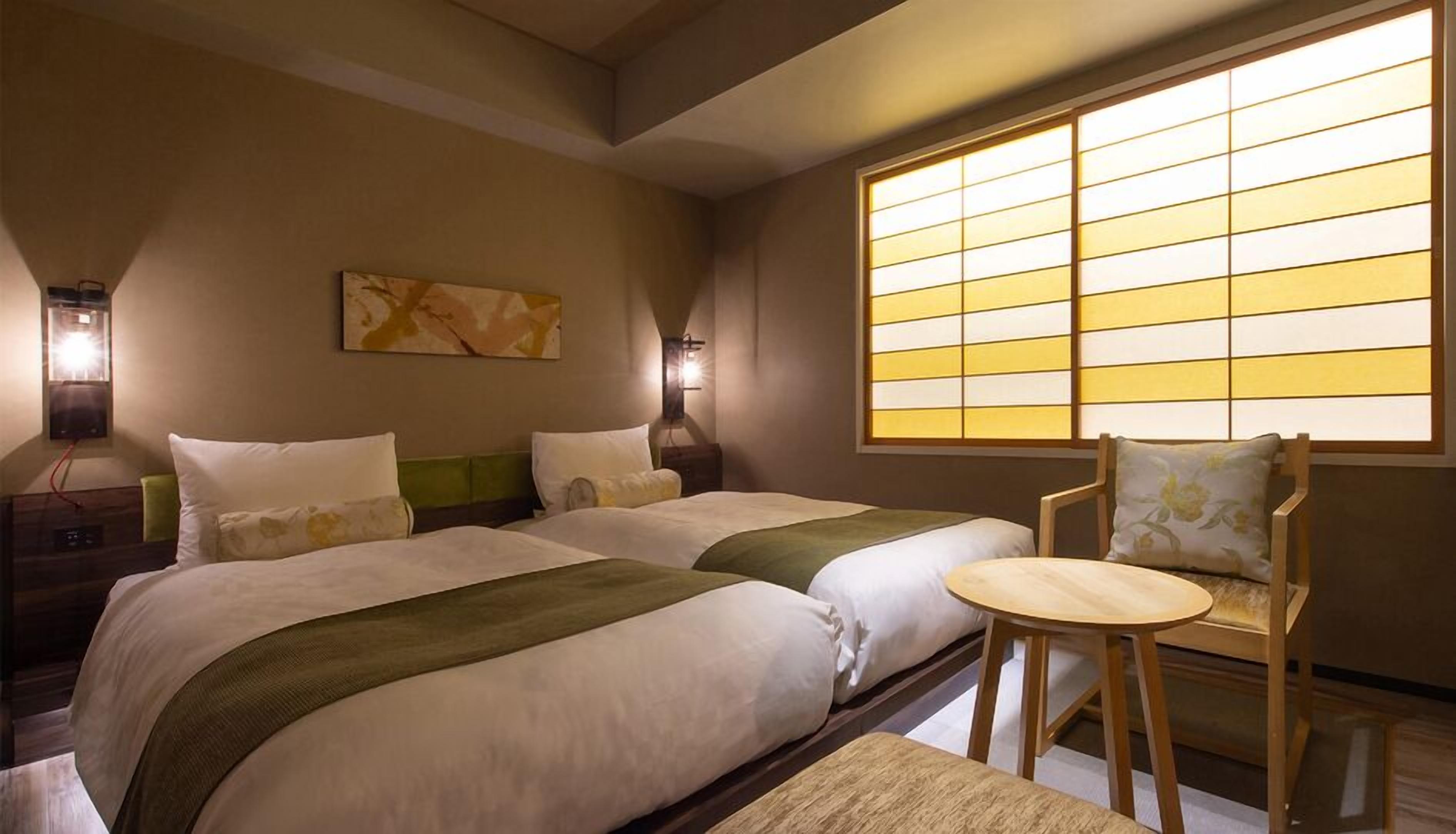 Quarto de hotel com duas camas, uma cadeira, uma mesa e uma janela grande com vista.