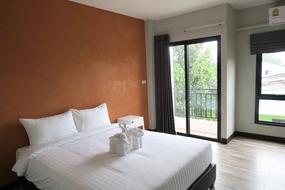 Habitación de hotel con una cama grande, una mesita de noche pequeña, una ventana con vistas a la vegetación y un balcón.