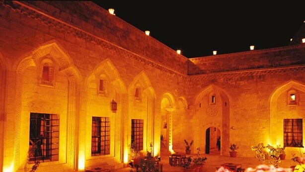 An outdoor courtyard with a brick wall, arched doorways, and potted plants. - Artuklu Kervansarayi - Special Class (Mardin)