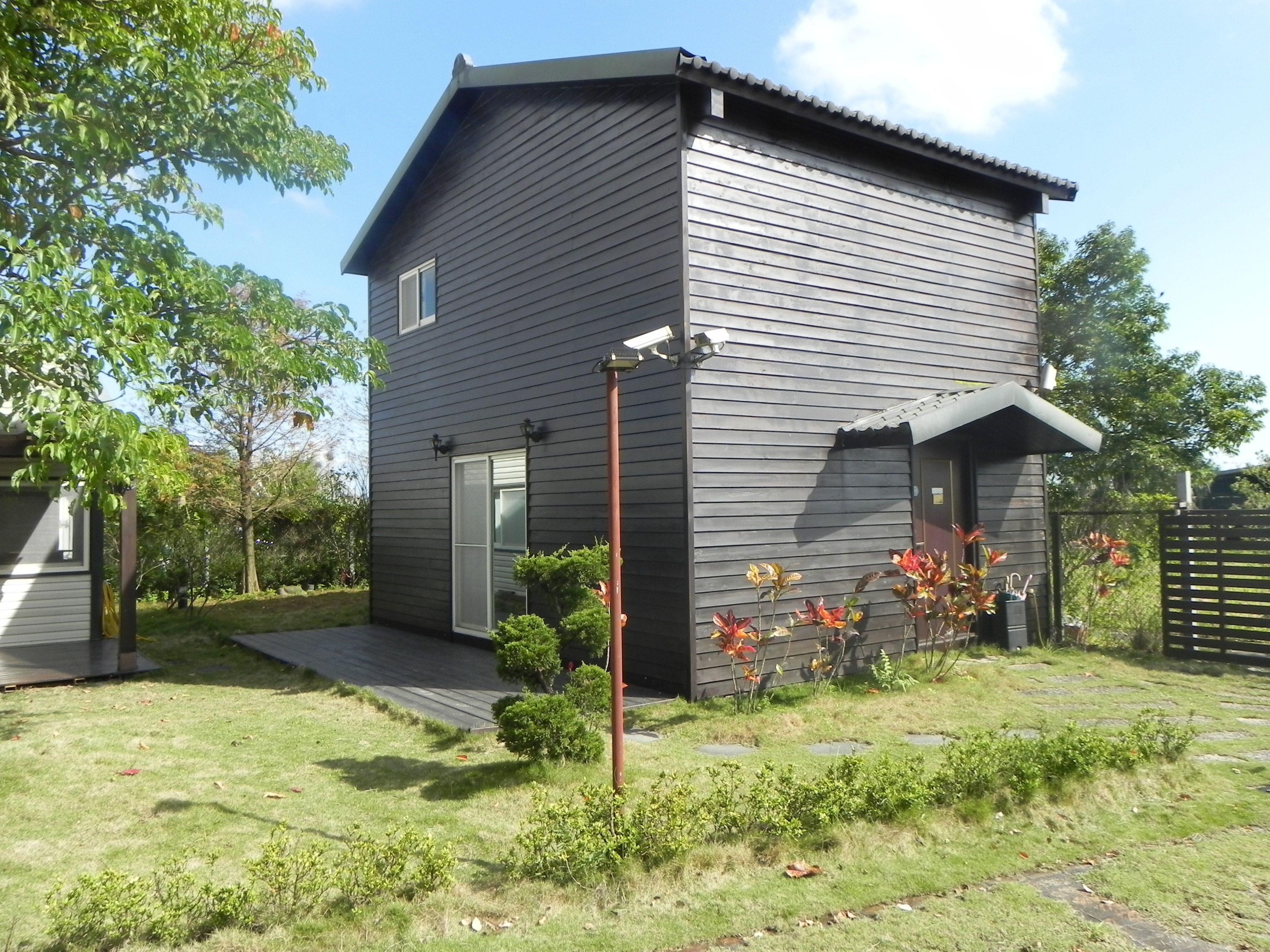 A modern two-story house with a dark exterior, a front porch, and a small garden with flowering plants.