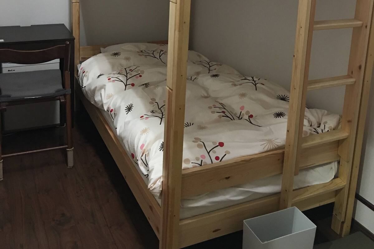 A bunk bed with a wooden frame, a desk with a chair, and a small trash bin.