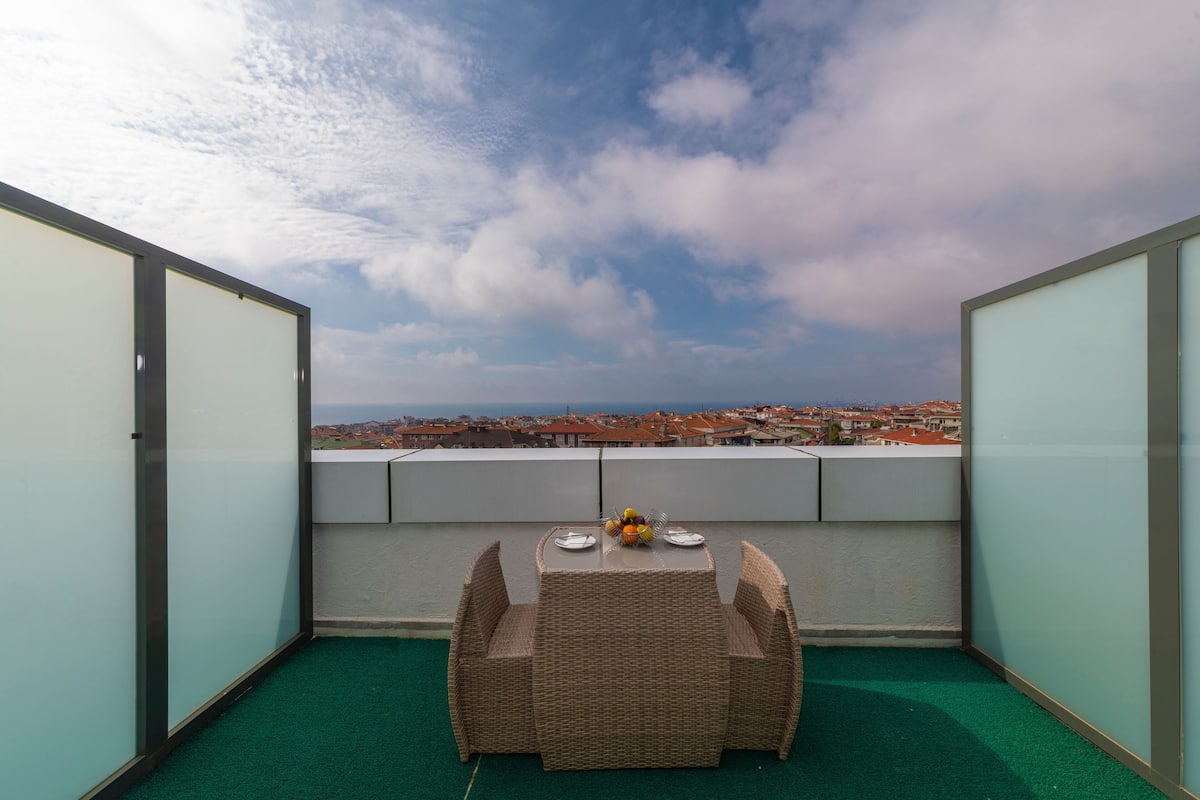 Un balcón con una mesa y sillas de mimbre, vista a un paisaje urbano y cielo despejado.