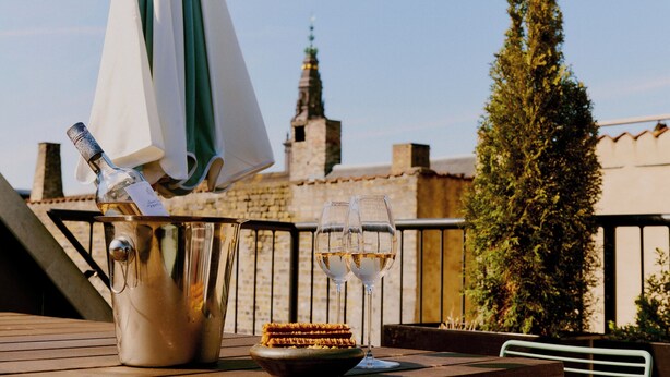 A table with a bottle of champagne, two glasses, and a plate of food, set against a backdrop of a building and a tree.