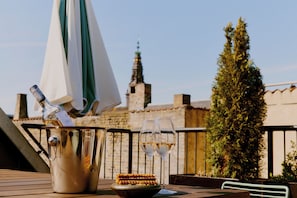 A table with a bottle of champagne, two glasses, and a plate of food, set against a backdrop of a building and a tree.