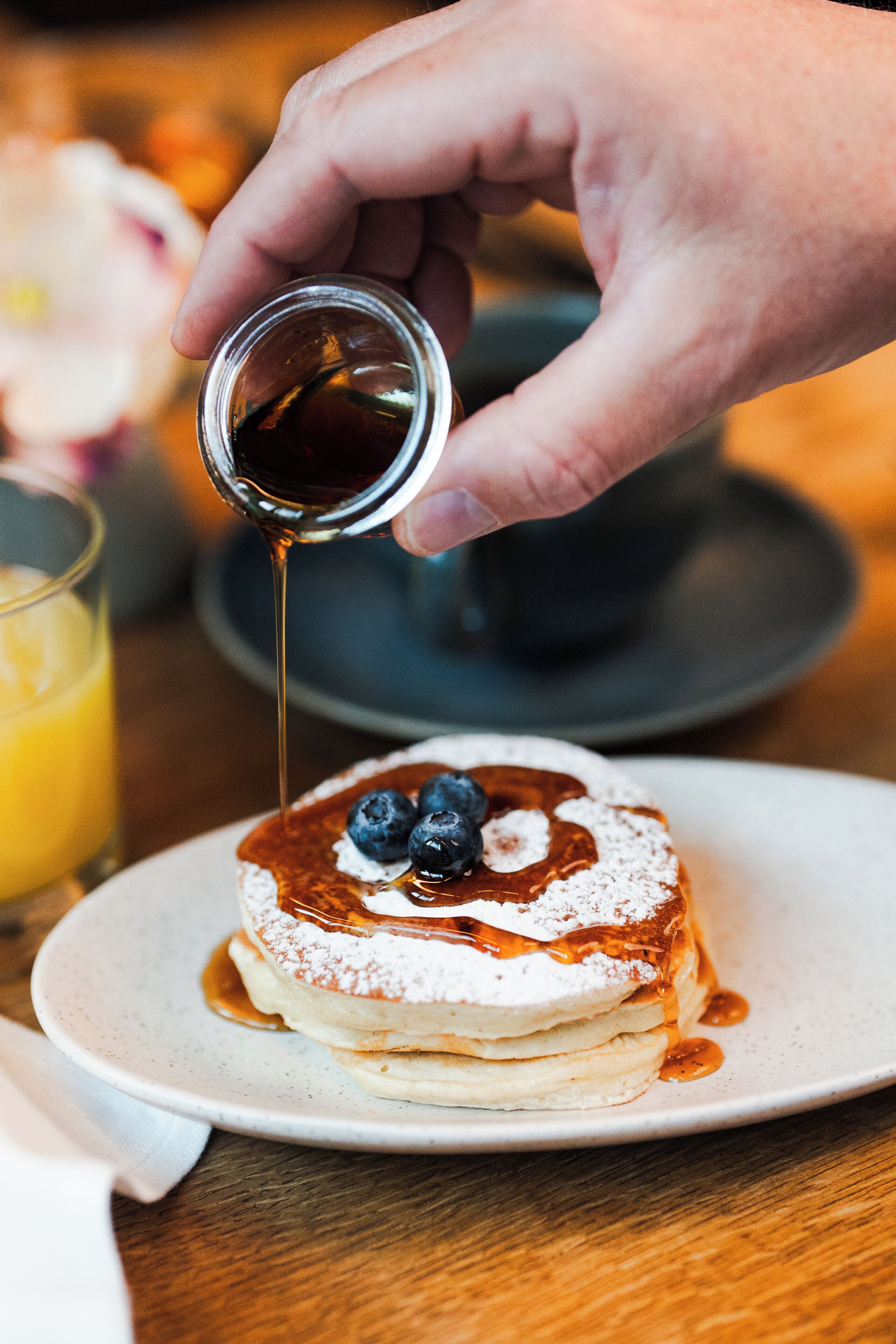 Eine Hand gießt Sirup über Pfannkuchen mit Blaubeeren auf einem Teller.