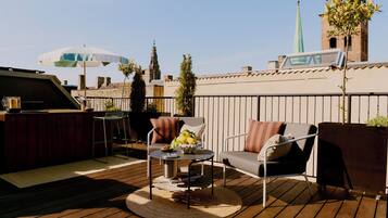 Een terras op het dak met buitenmeubels, een barbecue en uitzicht op een kerktoren.