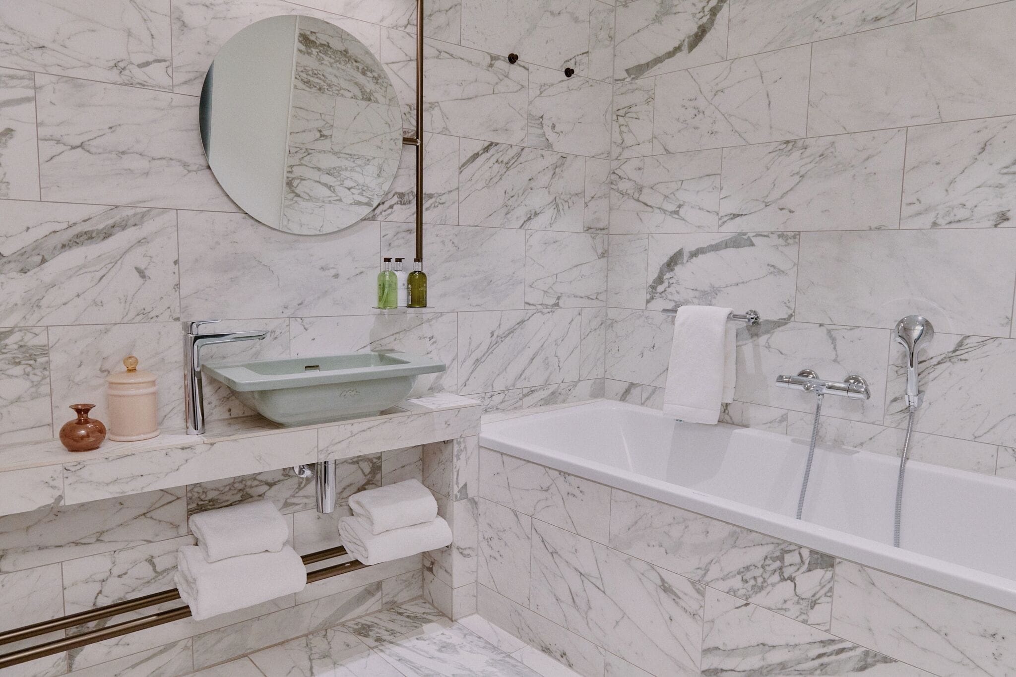 A bathroom with a marble-tiled wall, a round mirror, a sink, a bathtub, and a towel rack.