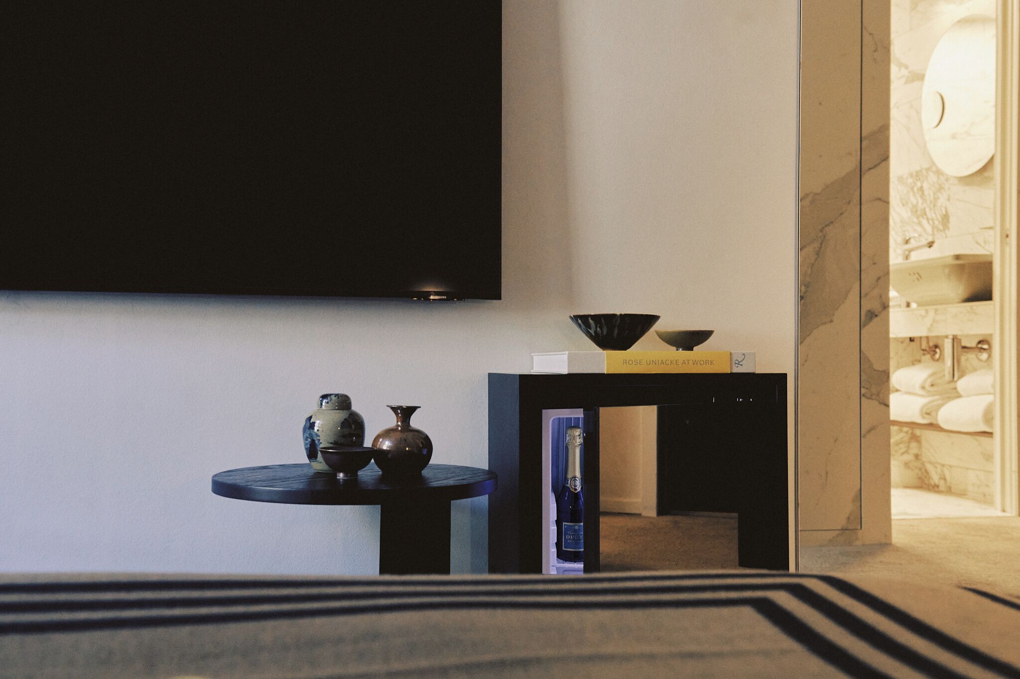 A modern hotel room with a flat-screen TV mounted on the wall, a small round table with decorative items, a bookshelf, and a marble bathroom visible through an open door.