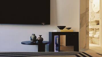A modern hotel room with a flat-screen TV mounted on the wall, a small round table with decorative items, a bookshelf, and a marble bathroom visible through an open door.