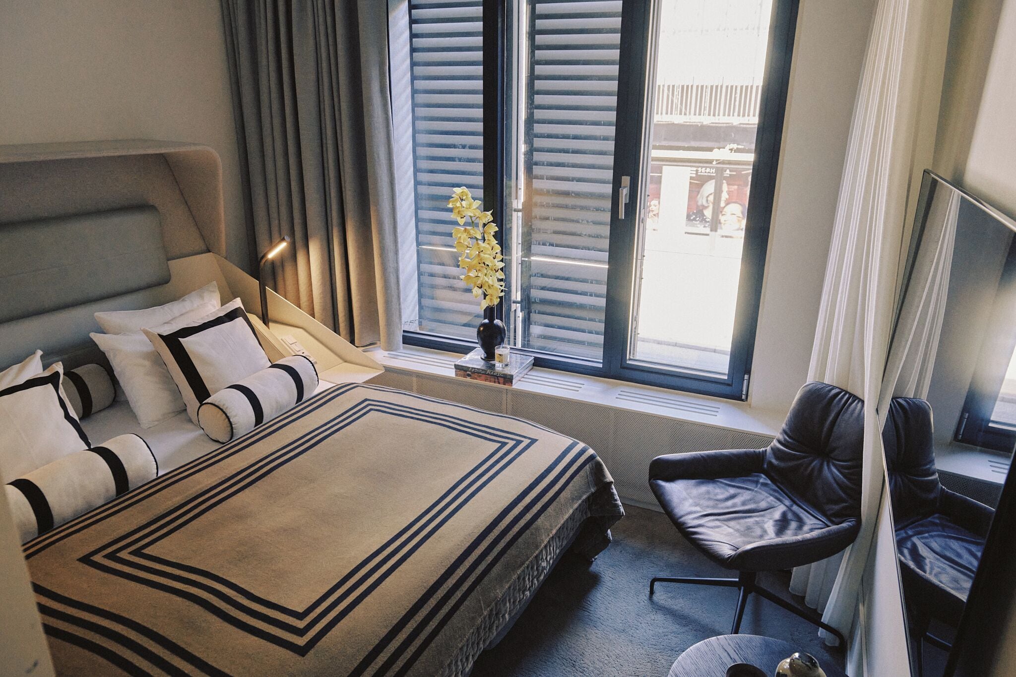 Habitación de hotel con cama, una silla, una mesita, un florero con flores y una ventana con persianas.