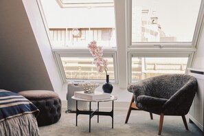 A cozy room with a view of a cityscape, a comfortable armchair, a small table with a vase of flowers, and a cushioned bench.