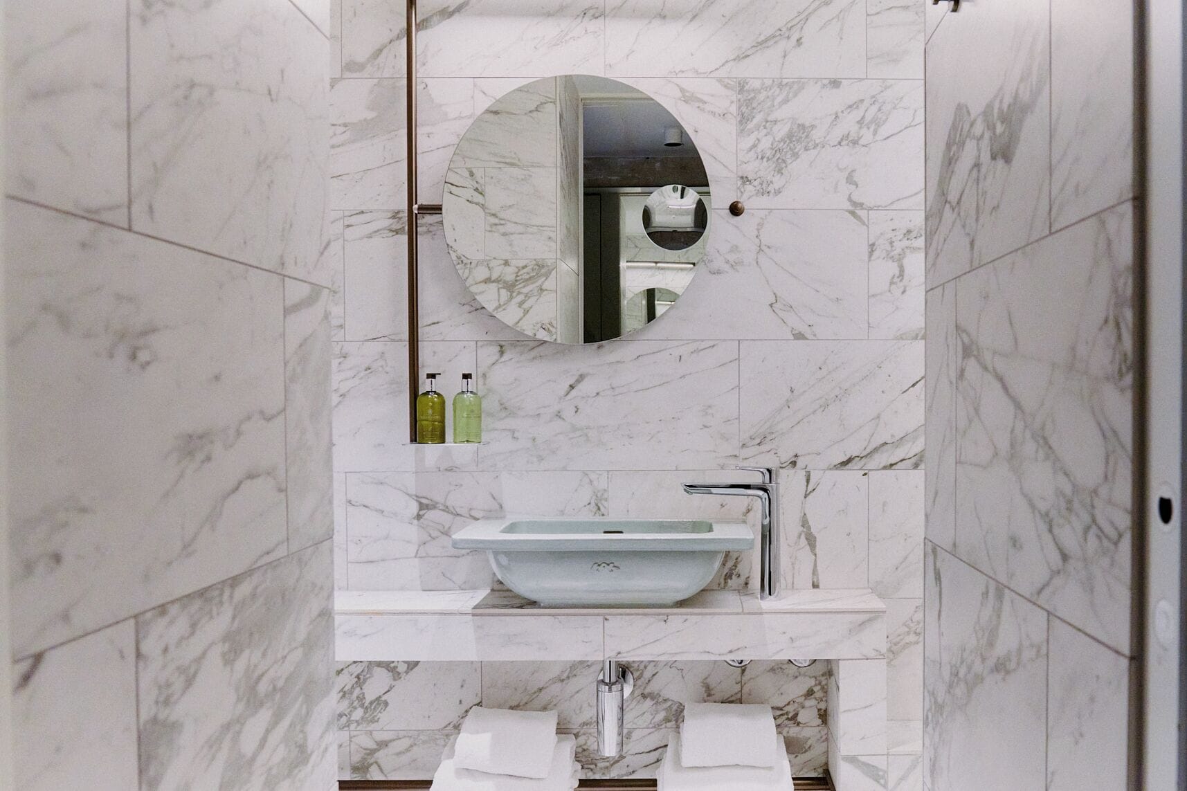 A bathroom with a marble wall, a round mirror, a vessel sink, and two bottles of liquid soap.