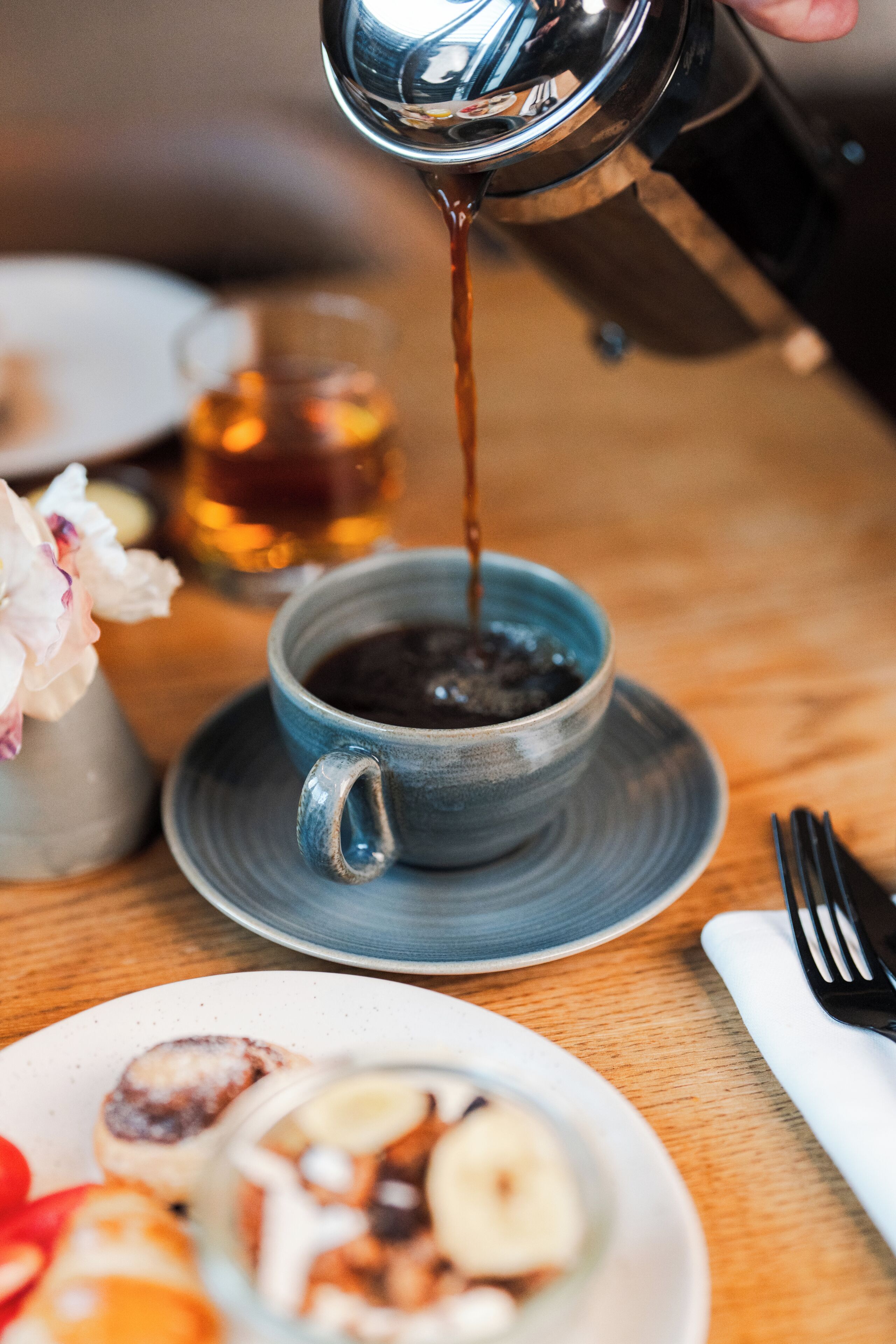 Une tasse de café est versée dans une tasse en céramique posée sur une soucoupe, avec une assiette de nourriture et un vase à fleurs en arrière-plan.