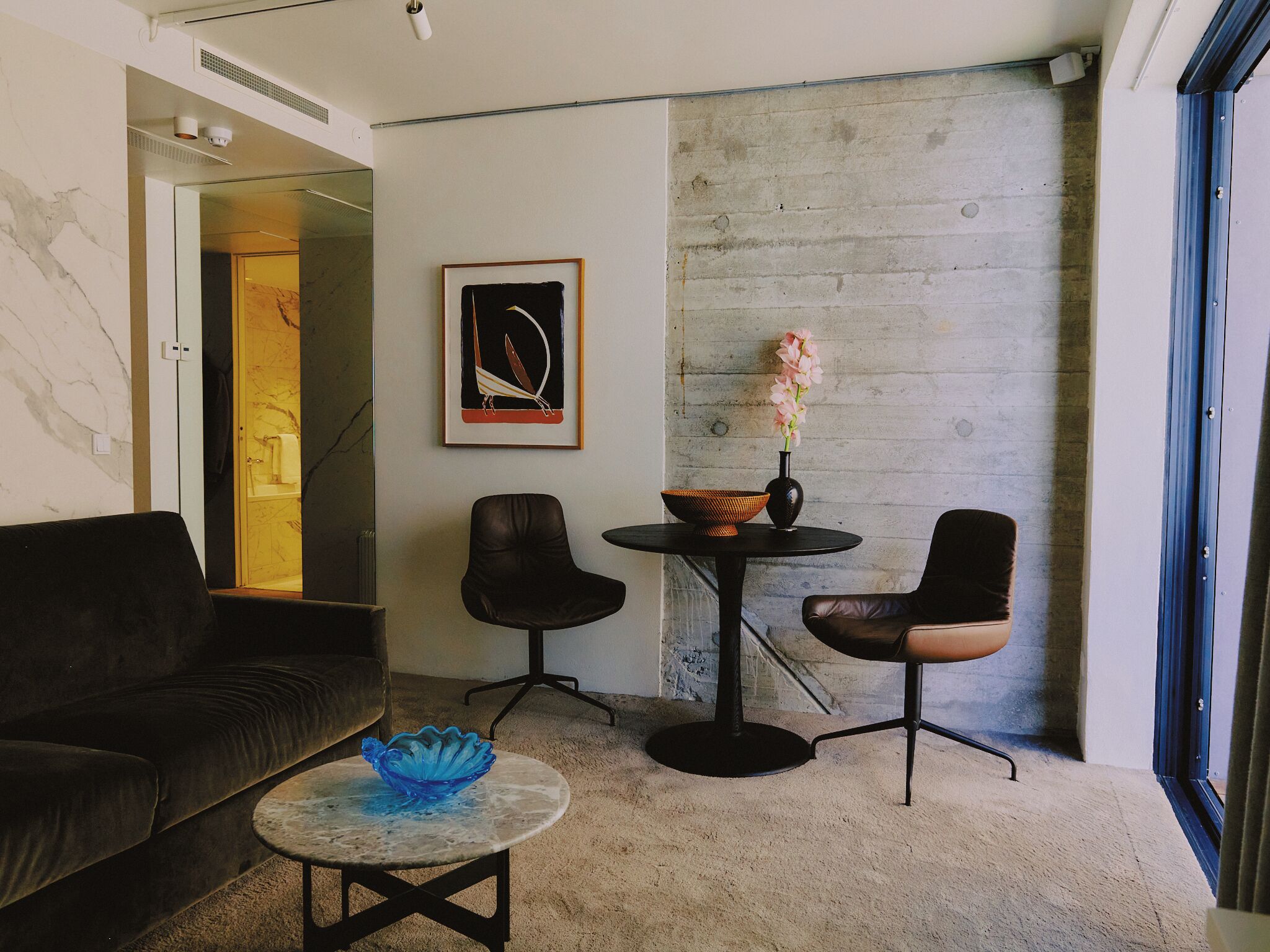 A modern living room with a marble wall, a dark leather sofa, a round coffee table with a blue bowl, two black chairs, a small round table with a vase, and a framed picture on the wall.