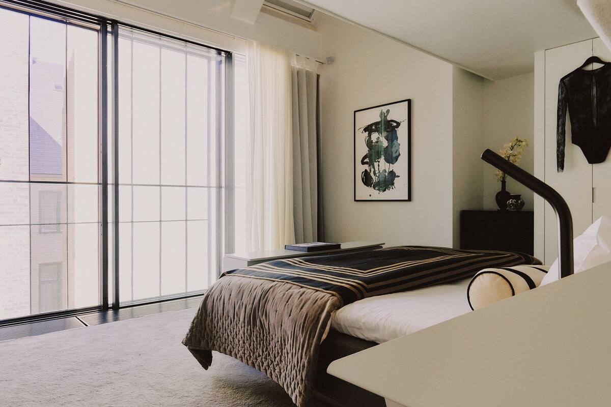 Ett hotellrum med ett stort fönster, en säng med ett randigt överkast, ett sängbord, ett inramat konstverk på väggen och ett hängande plagg.