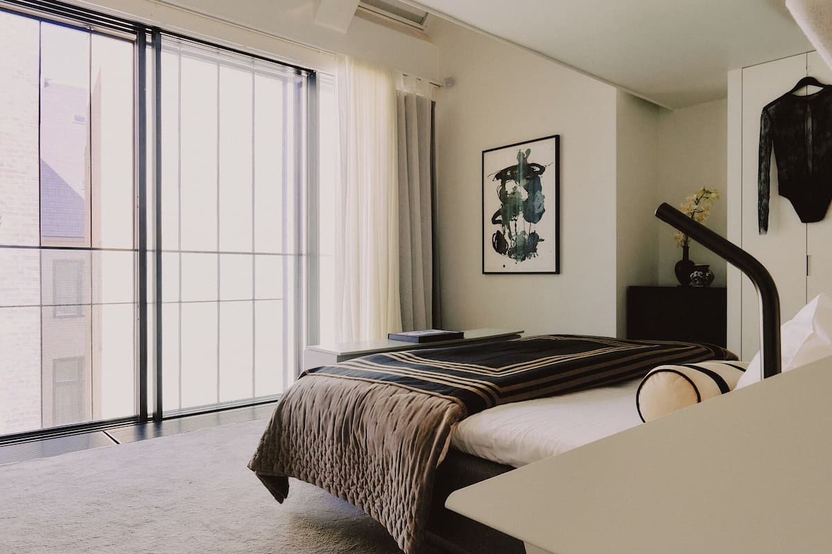 Een hotelkamer met een groot raam, een bed met een gestreept dekbed, een nachtkastje, een ingelijst kunstwerk aan de muur en een hangende kledingstuk.
