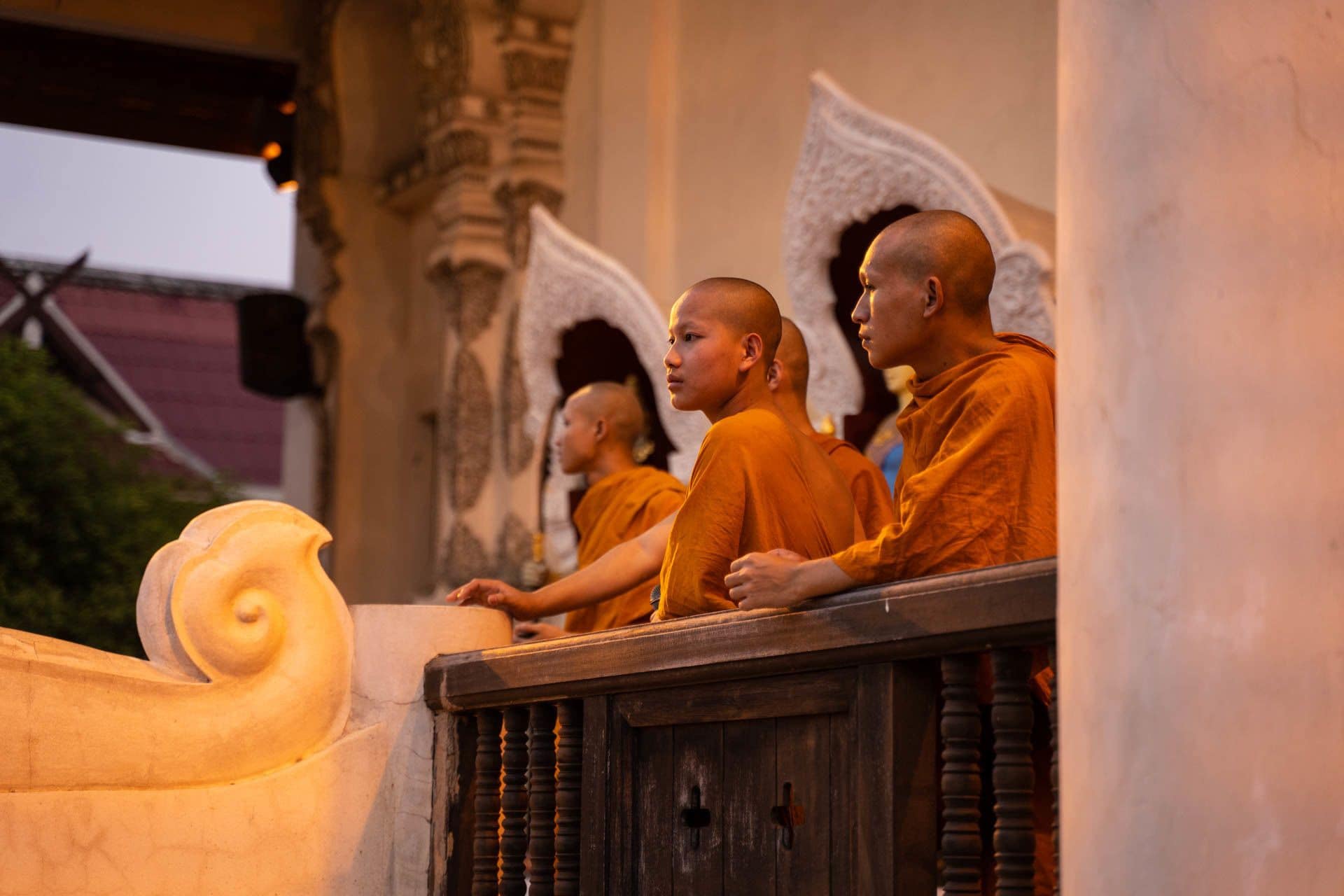 Monks in orange robes seated at a wooden lectern with a carved figure in front of them.