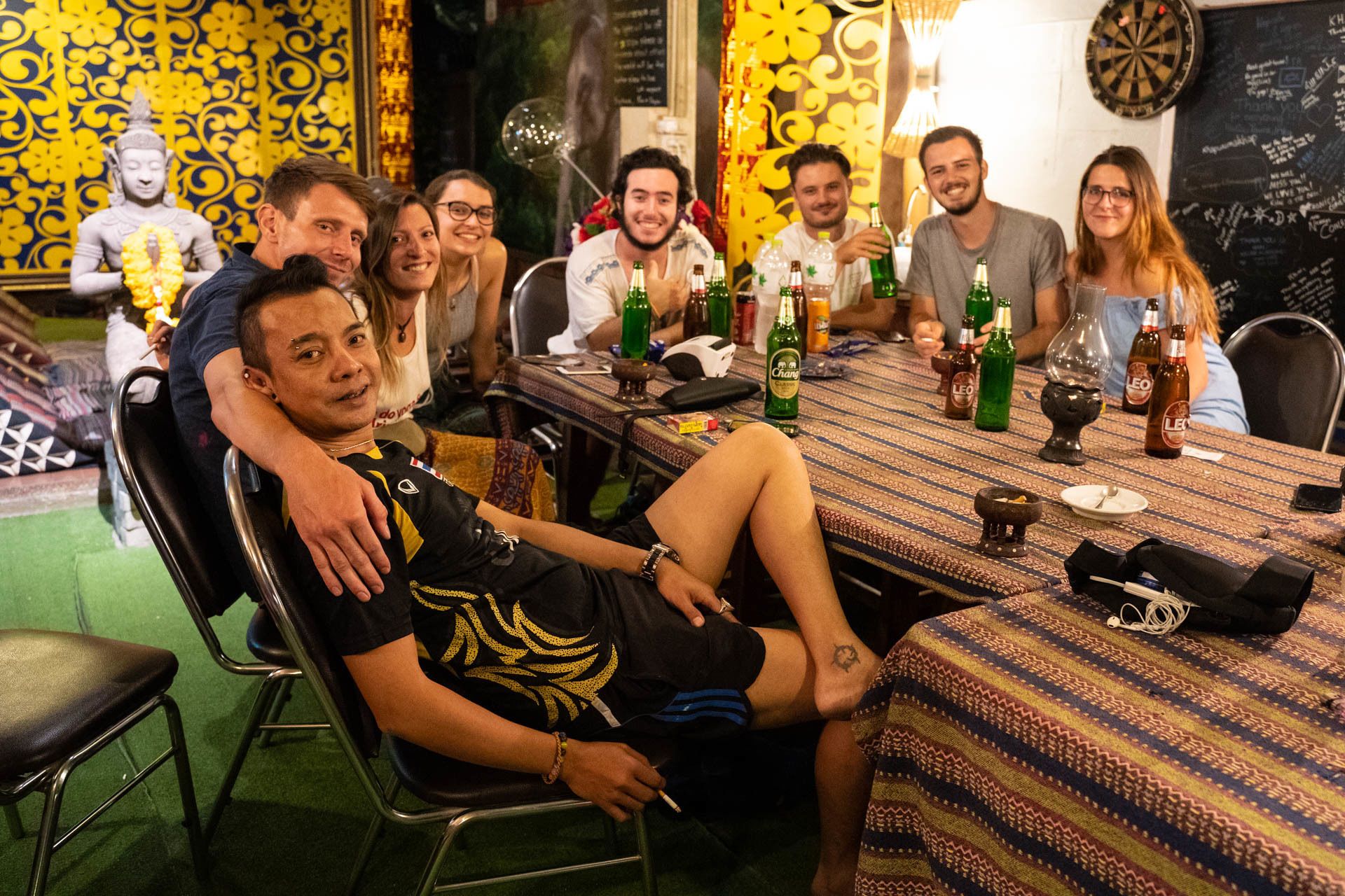 A group of people seated around a table with bottles and glasses, in a room with patterned wallpaper and a Buddha statue.