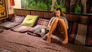 Un homme est assis sur un canapé, avec des coussins à motifs, une bouteille d’eau et une couverture.