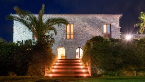A stone building with a staircase leading to the entrance, surrounded by greenery.