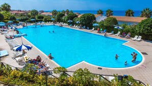 A large swimming pool with sun loungers and umbrellas, surrounded by palm trees and a view of the sea.