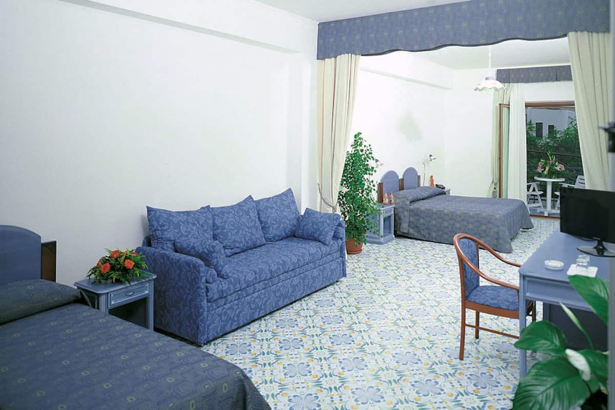 Ein Hotelzimmer mit einem blau gemusterten Sofa, einem Einzelbett, einem Schreibtisch mit Computer und einer Topfpflanze.