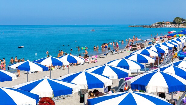 A beach with numerous blue and white striped umbrellas, people sunbathing, and a boat in the water.