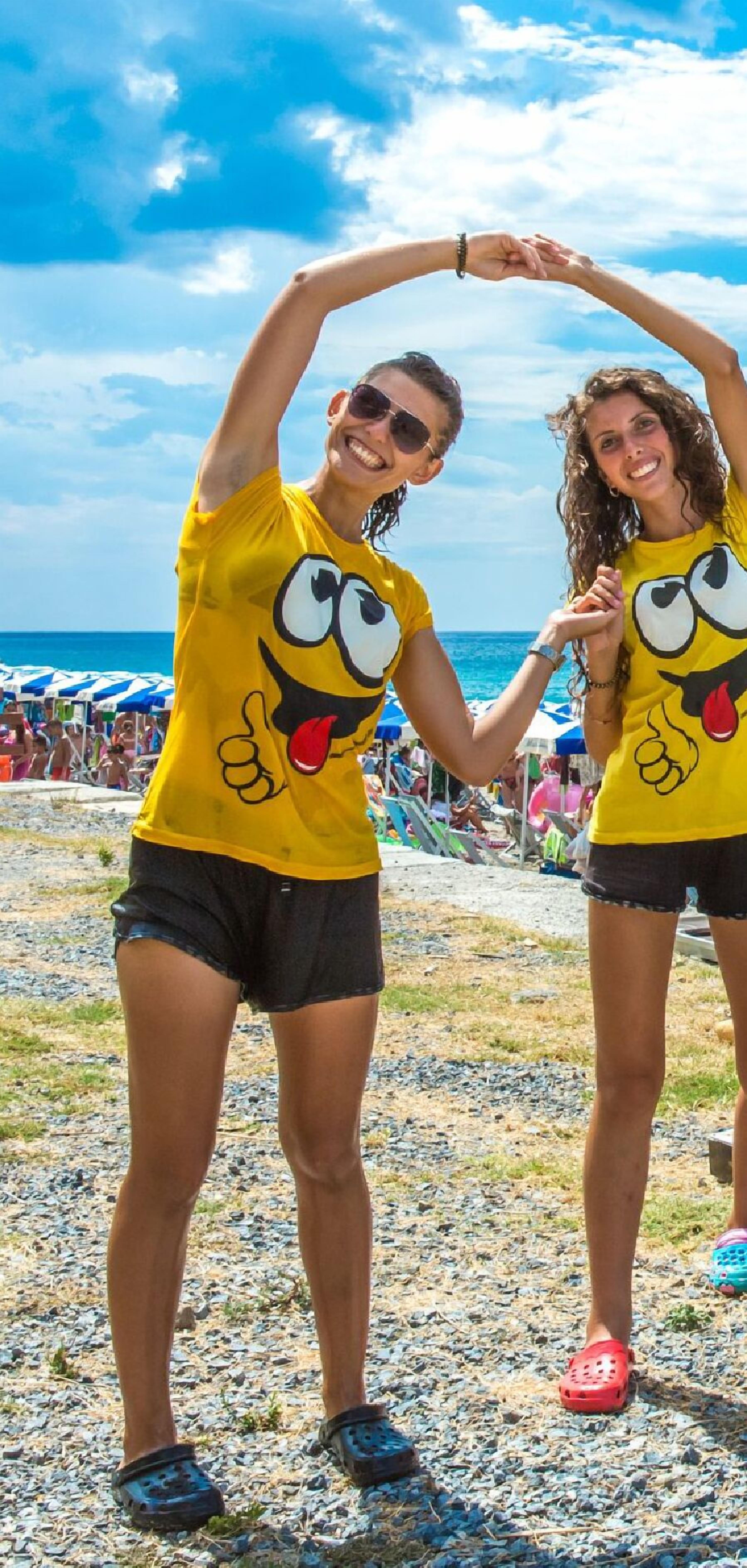 Due persone con magliette gialle raffigurate con un personaggio dei cartoni animati, in piedi su una spiaggia rocciosa con ombrelloni e altre persone sullo sfondo.