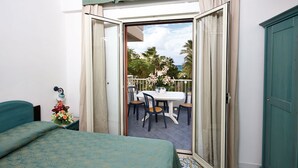 A hotel room with a bed, a balcony with a table and chairs, and a view of palm trees and the sea.