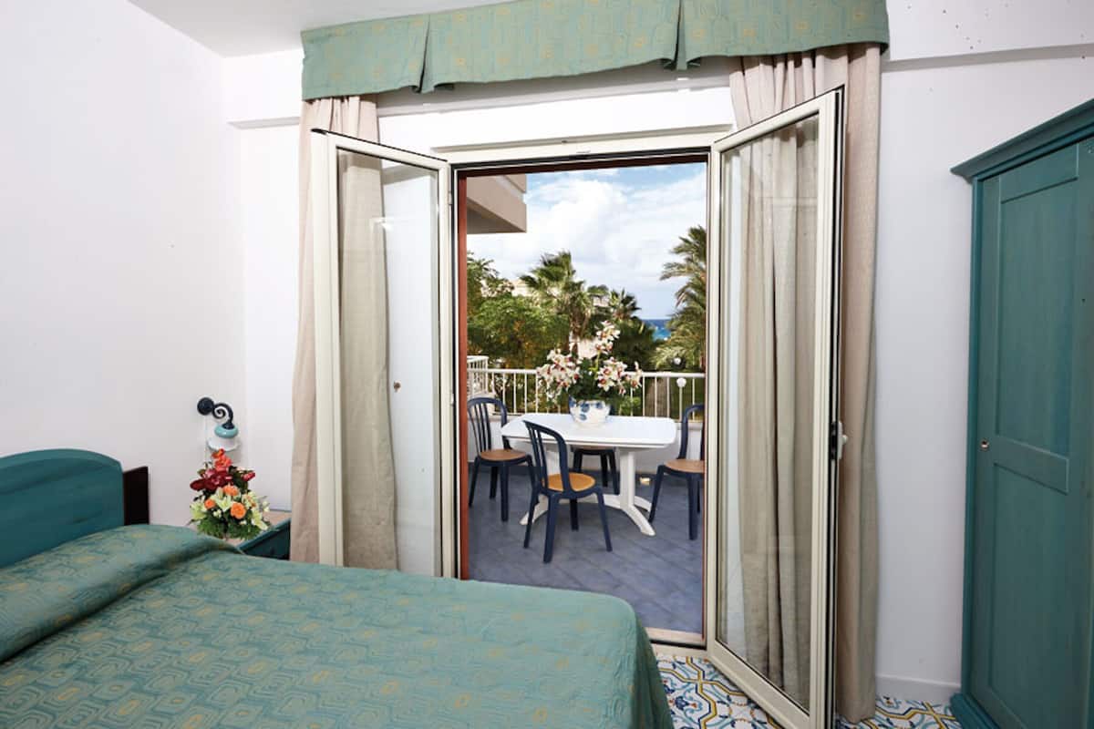 Ein Hotelzimmer mit Bett, einem Balkon mit Tisch und Stühlen sowie Blick auf Palmen und Meer.