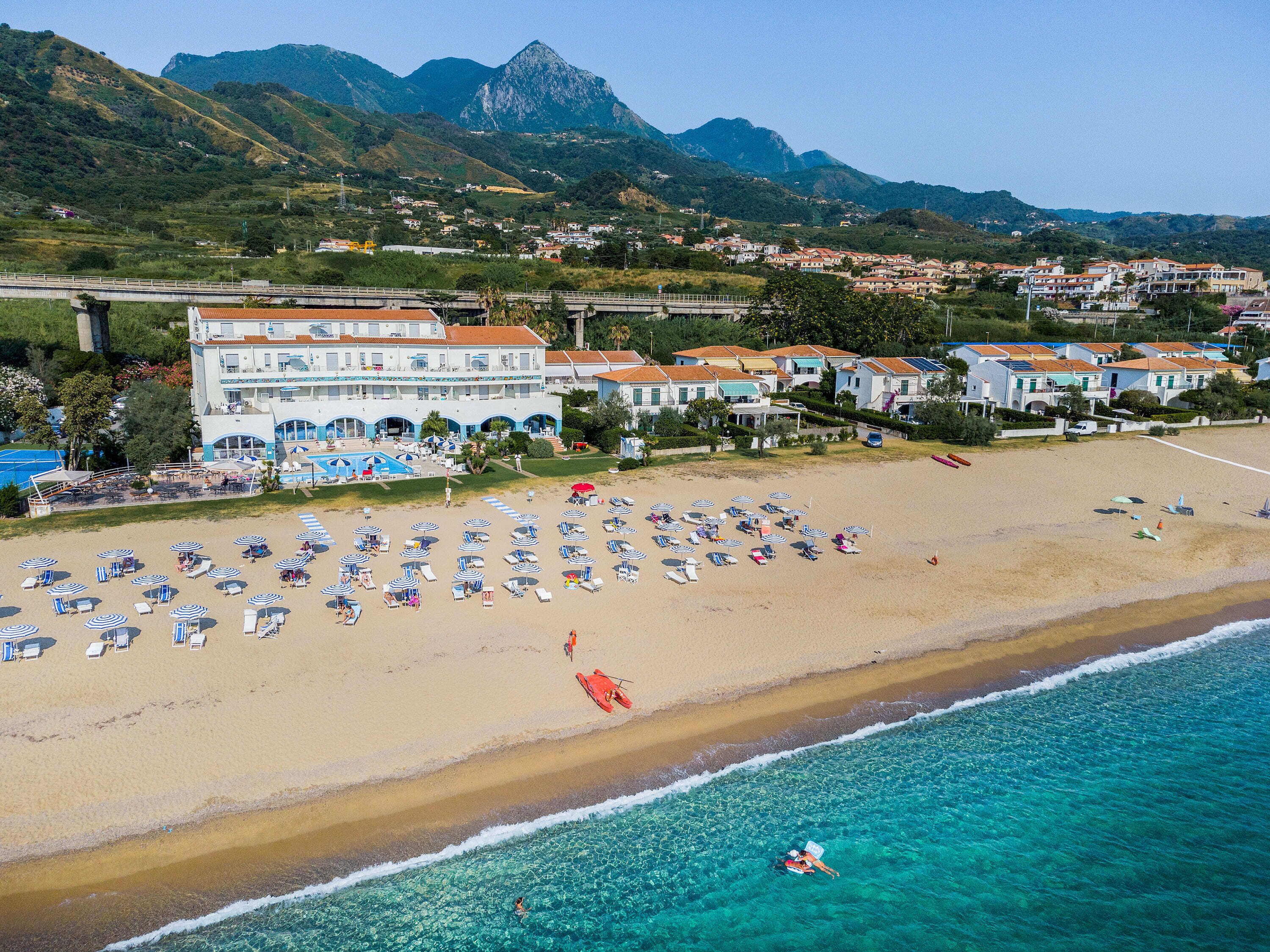 Un hôtel situé sur la côte, avec une plage, une eau cristalline et un paysage montagneux en arrière-plan.