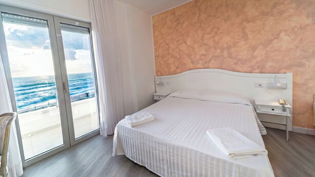 A hotel room with a large bed, a nightstand, a wall-mounted lamp, and a view of the ocean through a sliding door. - Hotel Poseidon (Belvedere Marittimo)