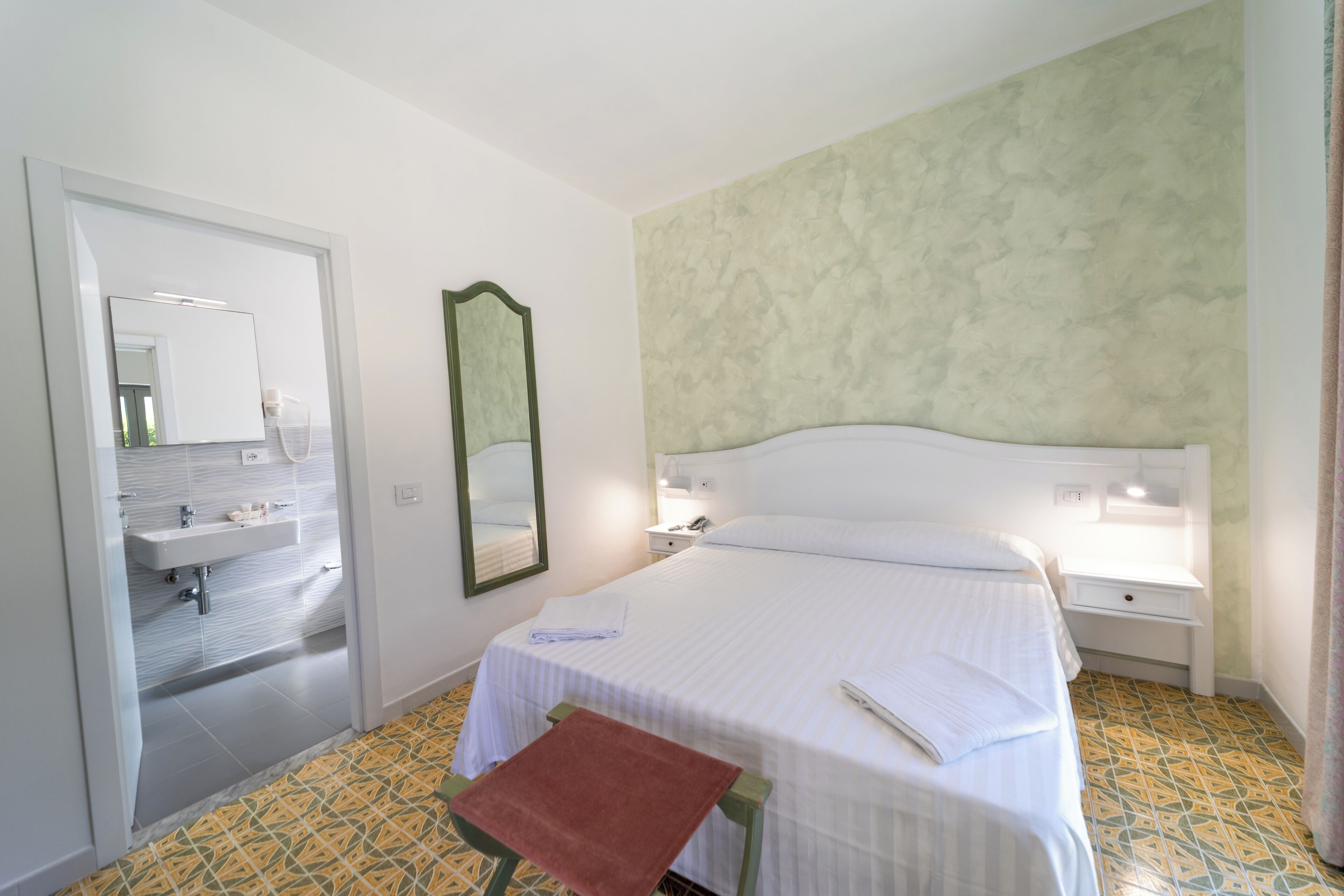 Une chambre d’hôtel comprenant un lit, une table de chevet, une commode, un miroir et une salle de bain avec un lavabo et un miroir.