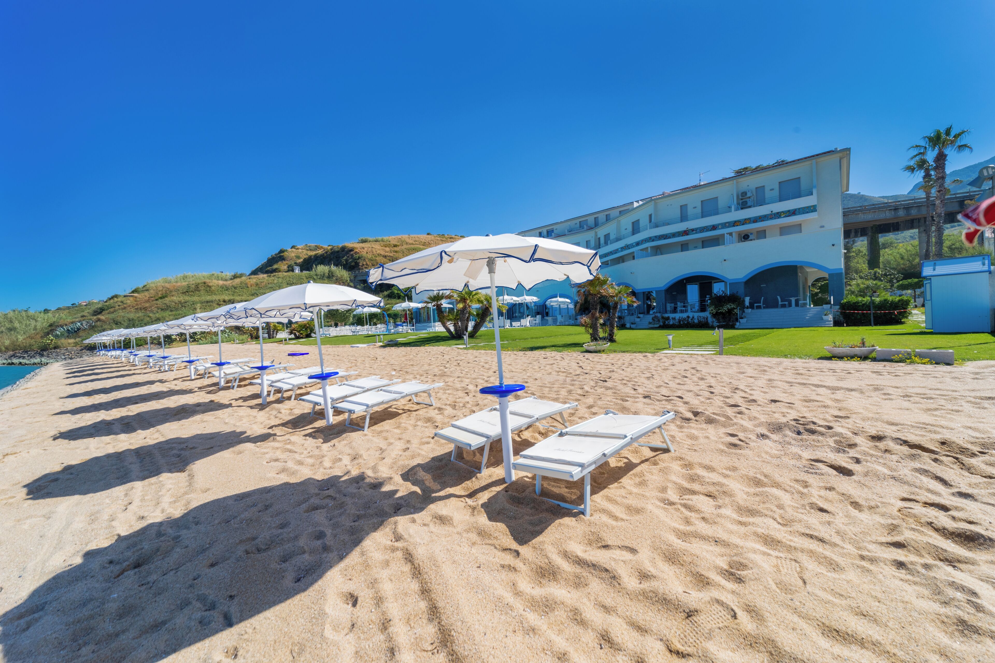 Une plage avec des chaises longues et des parasols, un bâtiment doté d’un balcon et un ciel bleu dégagé.