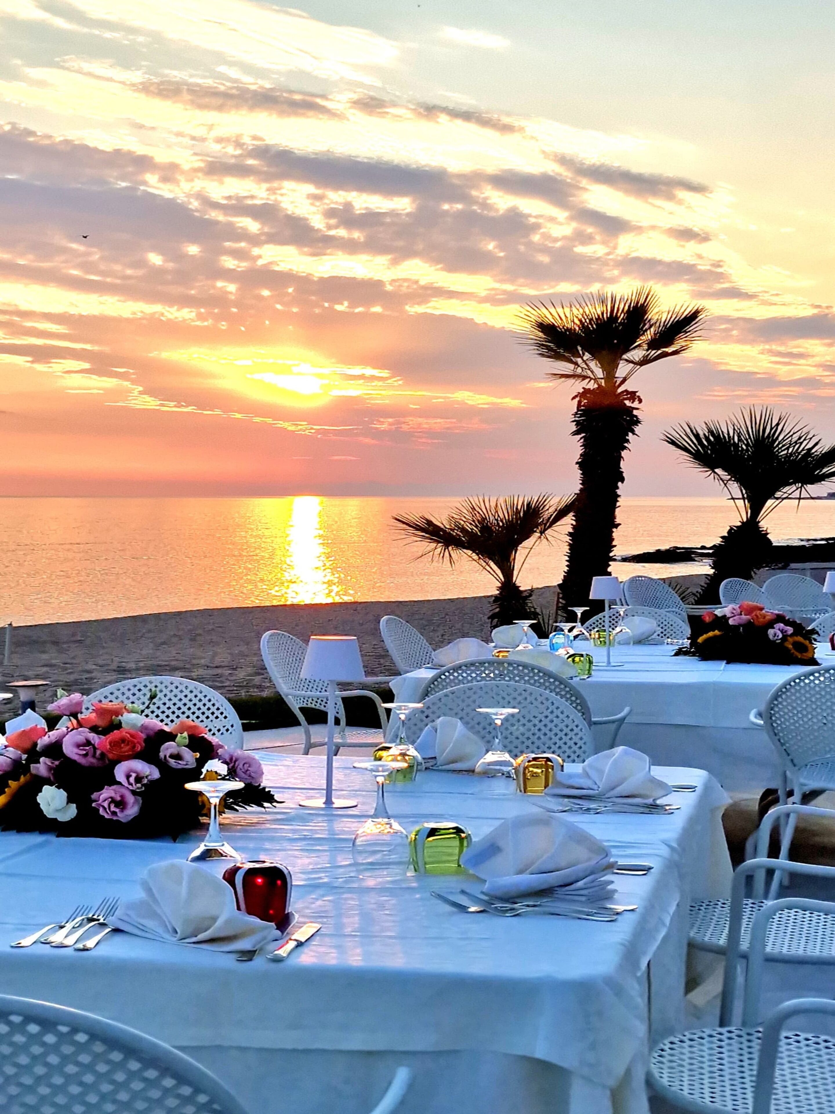 Une vue sur le coucher de soleil, avec des tables dressées pour le dîner, des palmiers et une plage en arrière-plan.