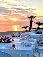 Vista al atardecer con mesas preparadas para cenar, palmeras y una playa al fondo.