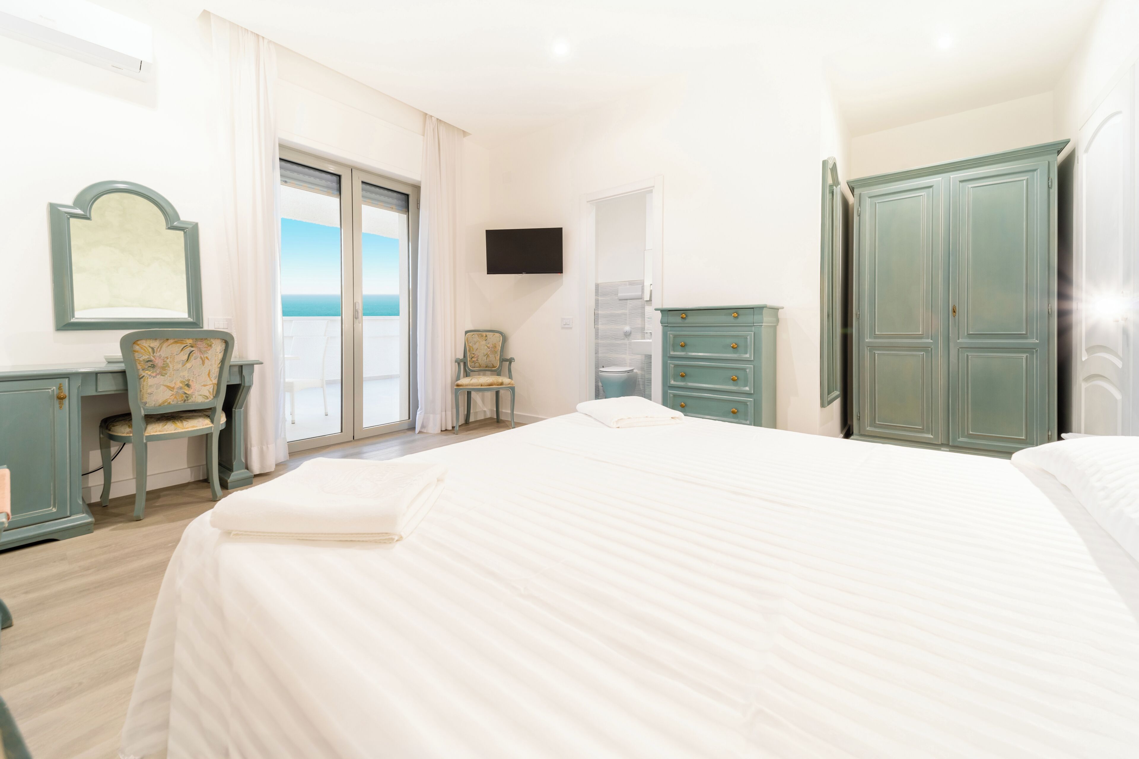 A bedroom with a bed, a chair, a dresser, a television, and a view of the sea.