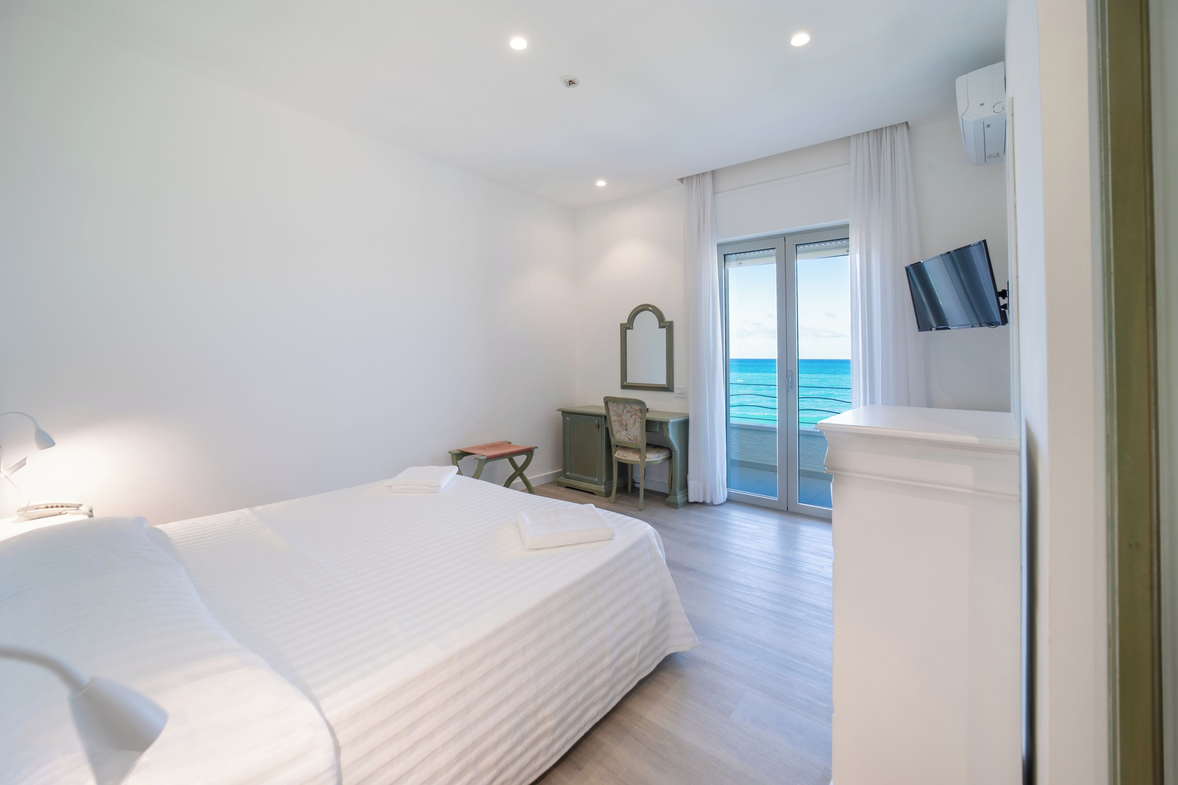 A hotel room with a bed, a desk, a chair, a TV, and a view of the sea.