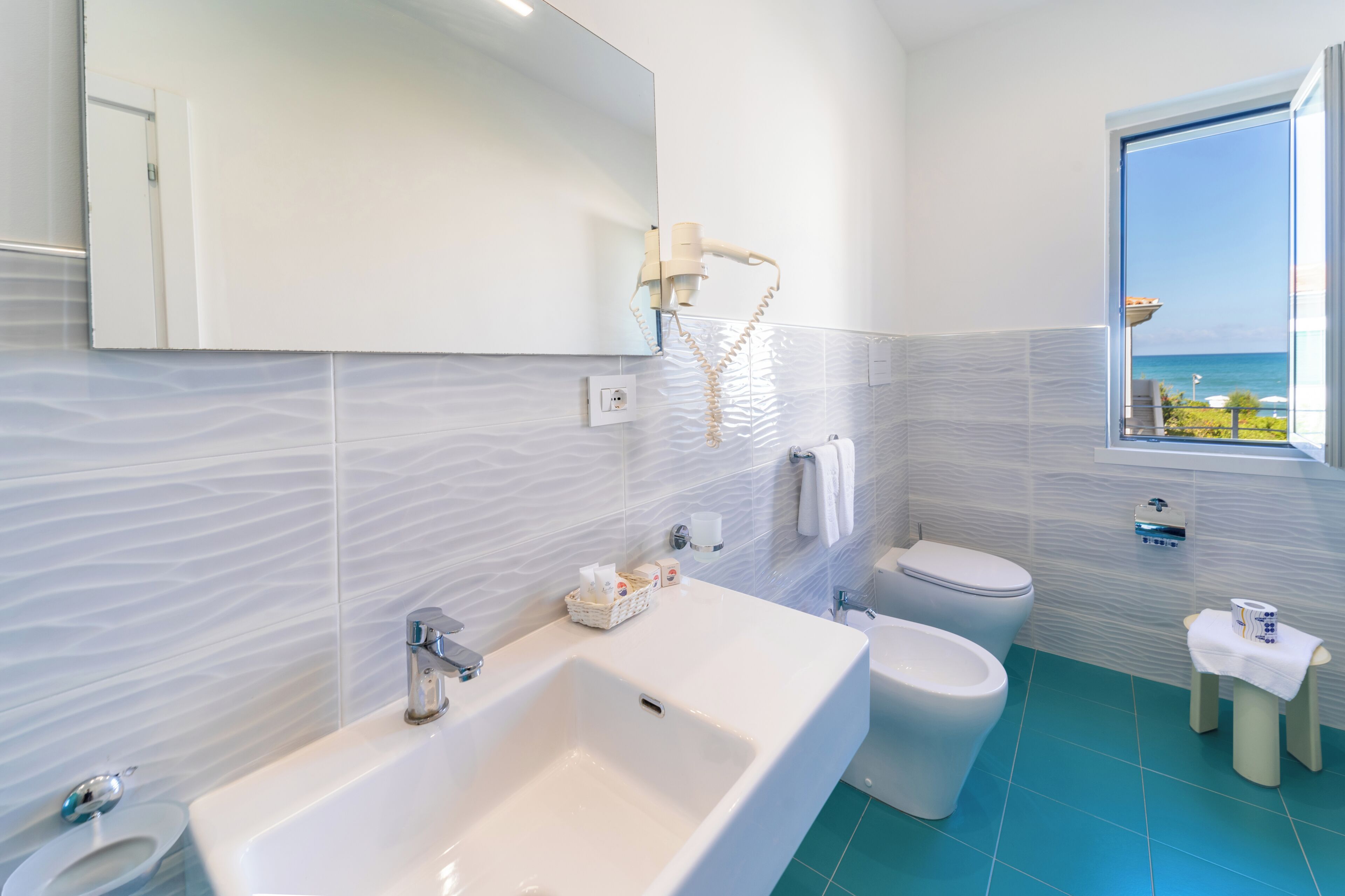 A modern bathroom with a large sink, toilet, and bidet, a mirror, and a window with a view of the sea.