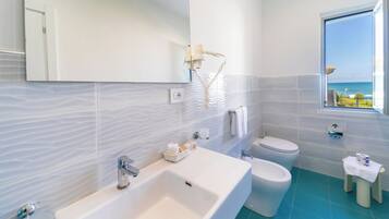 A modern bathroom with a large sink, toilet, and bidet, a mirror, and a window with a view of the sea.