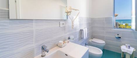 Een moderne badkamer met een groot wastafel, toilet en bidet, een spiegel, en een raam met uitzicht op zee.