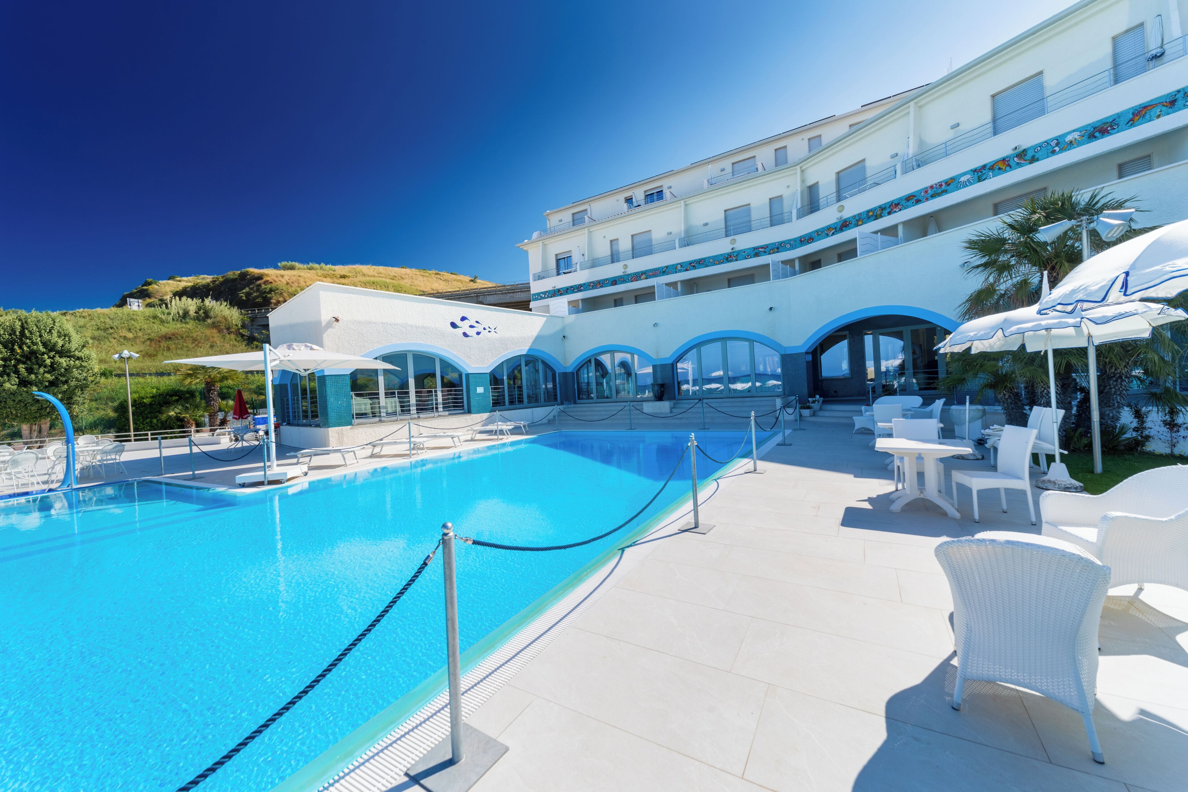Una piscina con agua azul cristalina, rodeada de sillas de descanso y sombrillas blancas, y un edificio con una decoración azul y blanca al fondo.
