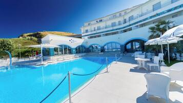 Una piscina con agua azul cristalina, rodeada de sillas de descanso y sombrillas blancas, y un edificio con una decoración azul y blanca al fondo.