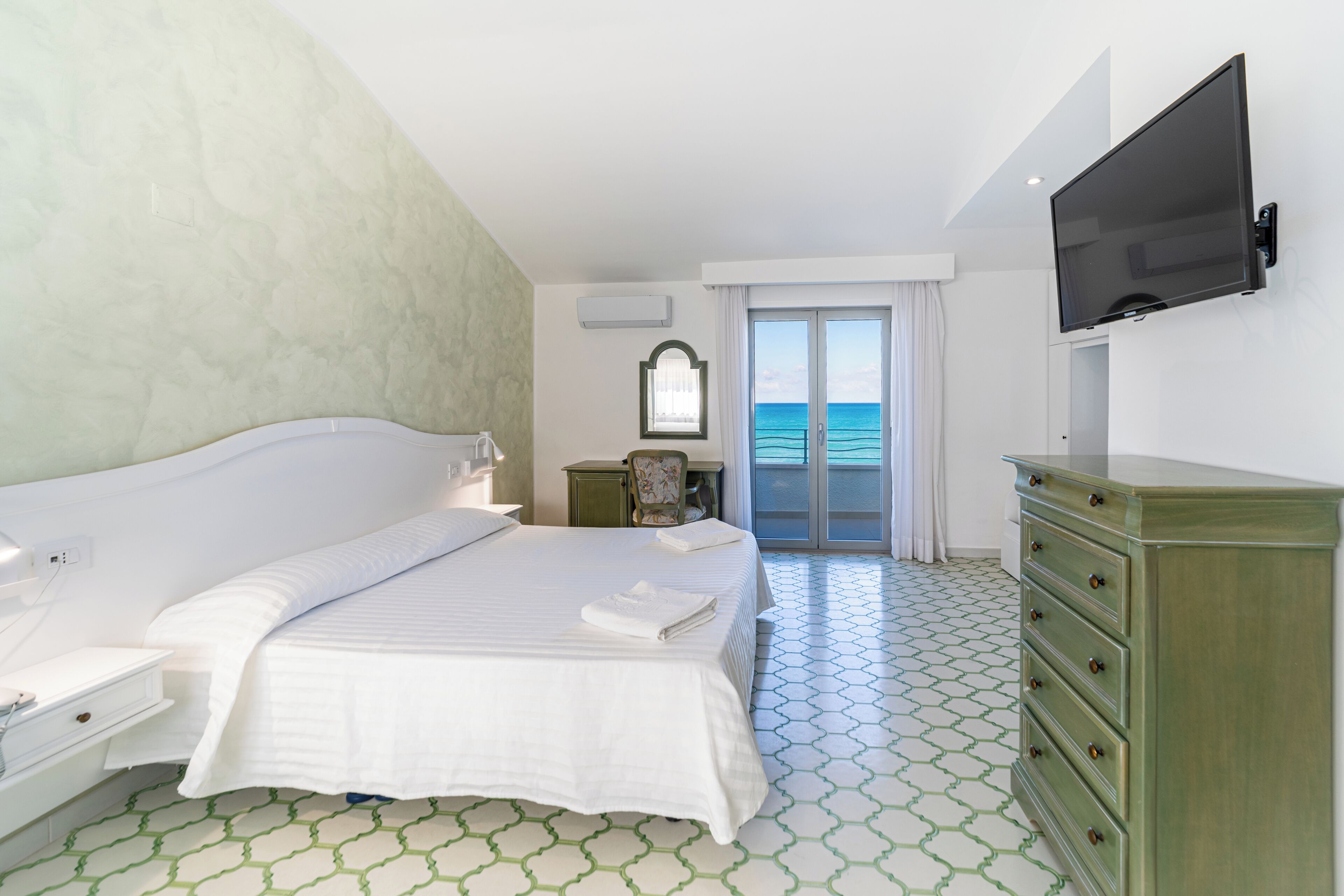 A hotel room with a bed, a dresser, a television, a window with a view of the sea, and a patterned floor.