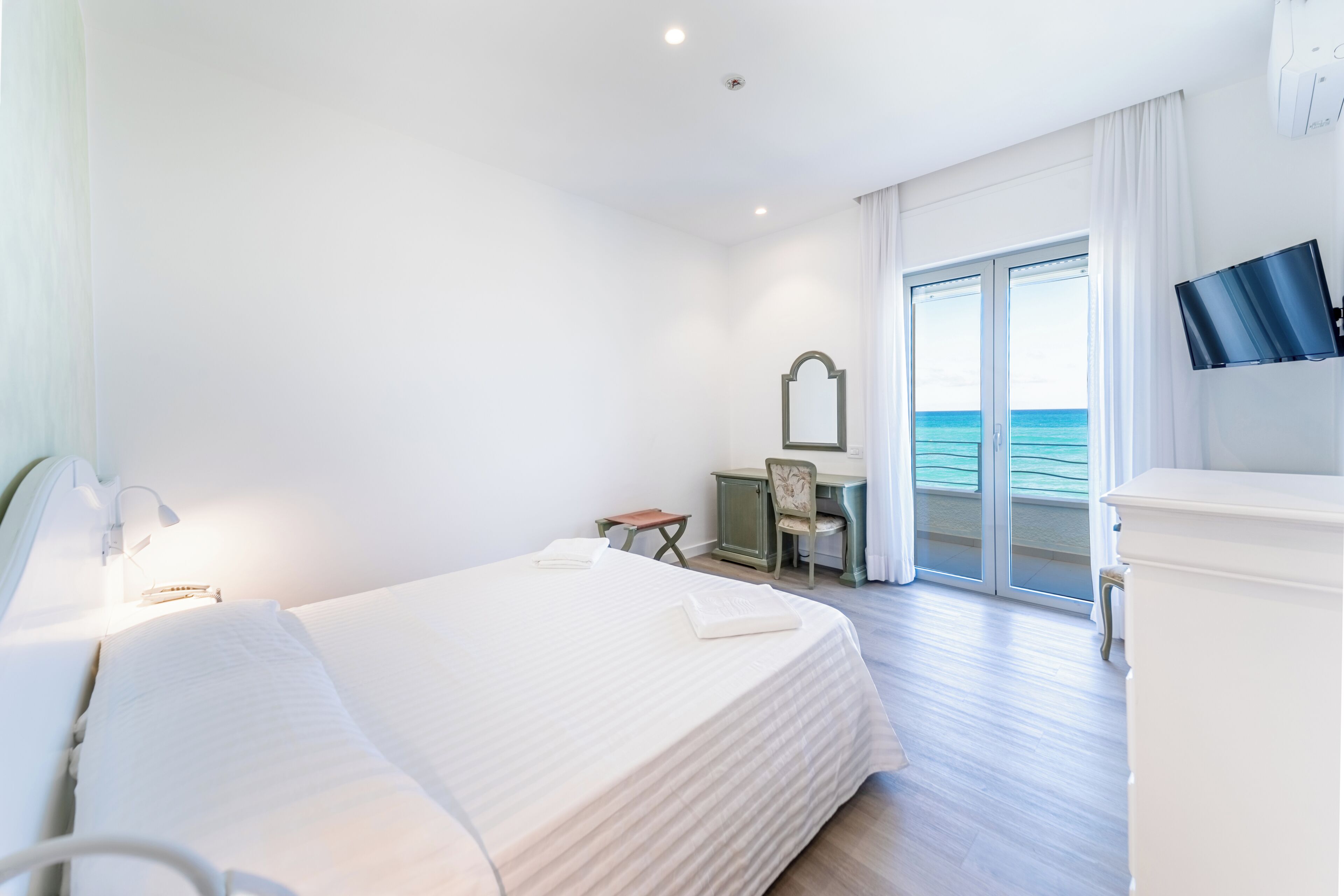 Una camera d'albergo con un letto, una scrivania, una sedia, una televisione e vista mare attraverso la porta scorrevole in vetro.