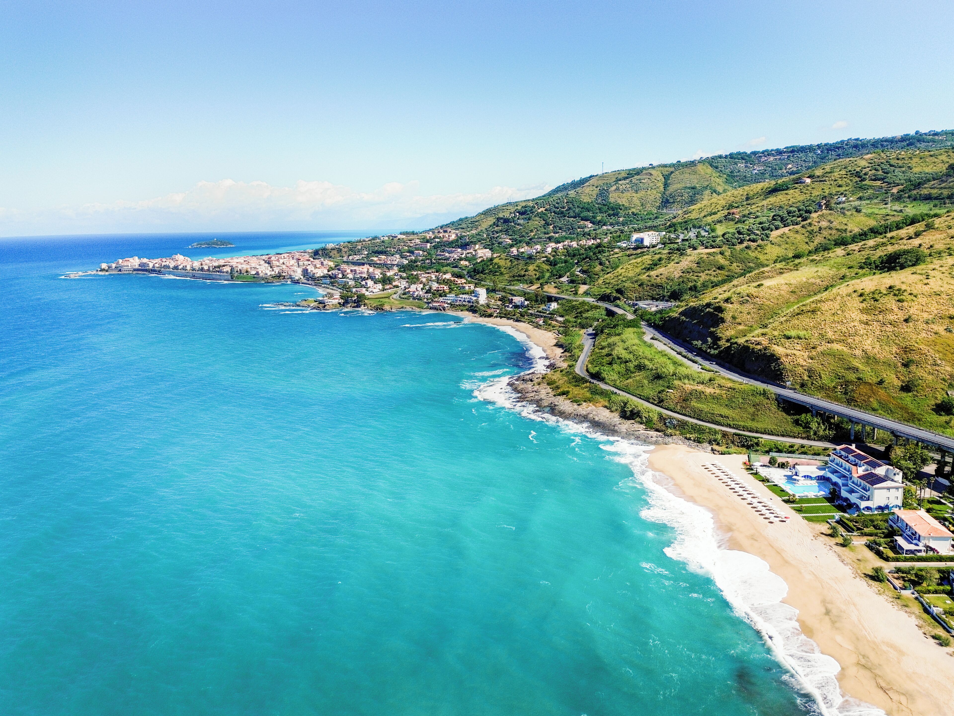 Una località costiera con una spiaggia, una strada che serpeggia lungo la costa e un paesaggio collinare sullo sfondo.
