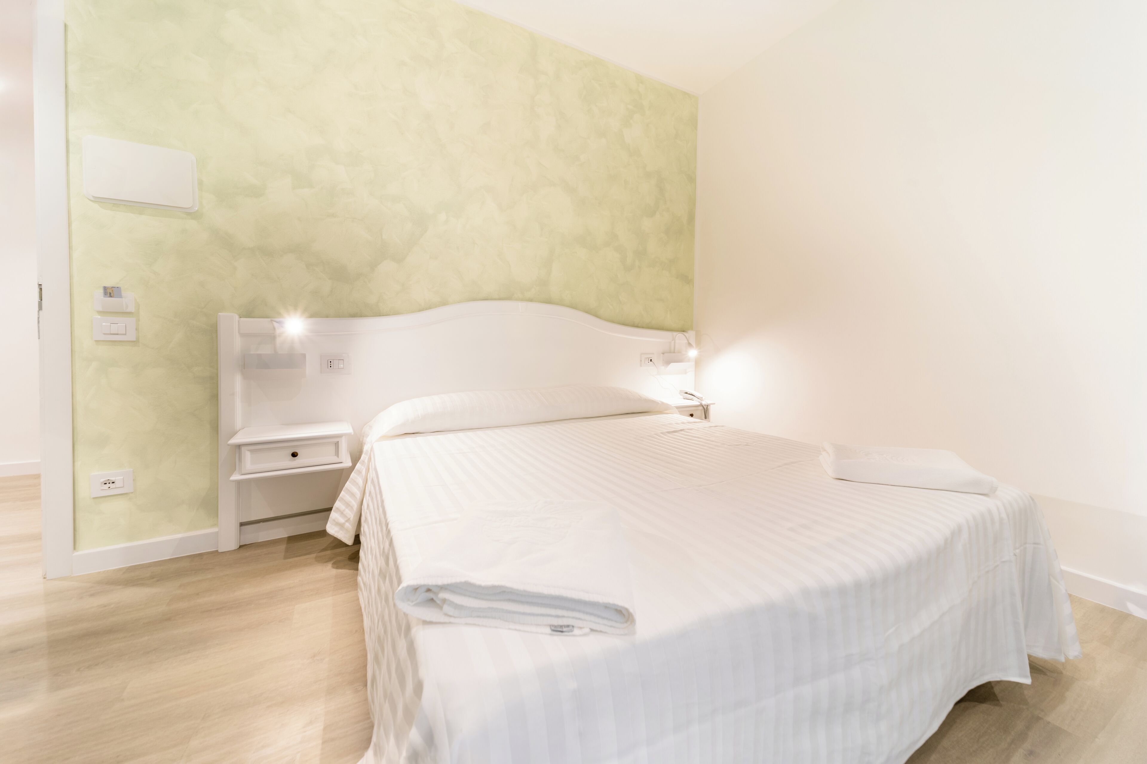 A hotel room with a bed, bedside tables, and a light-colored wall.