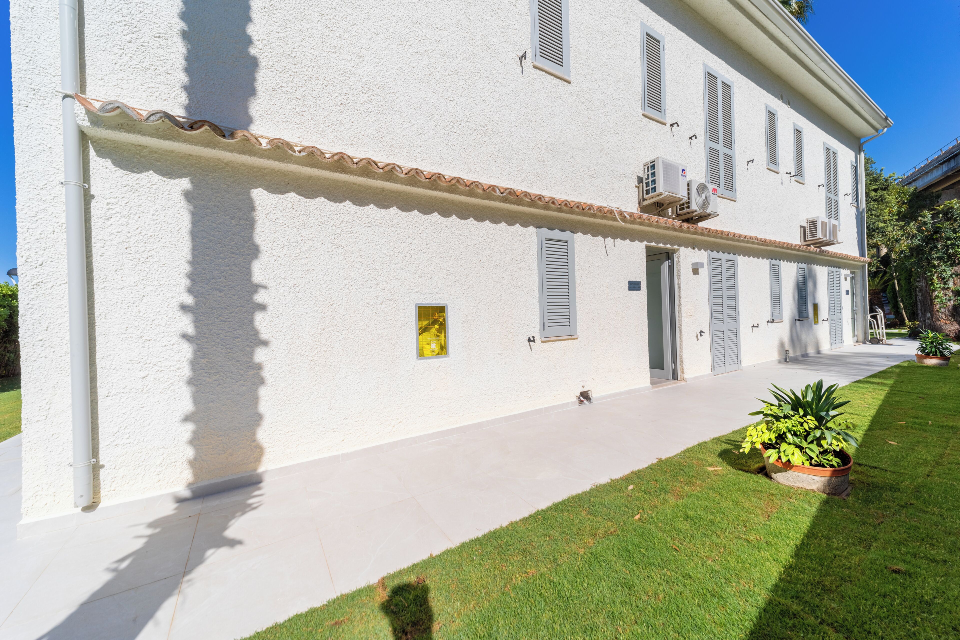 A white building with a tiled roof, a paved walkway, and a small garden with plants.