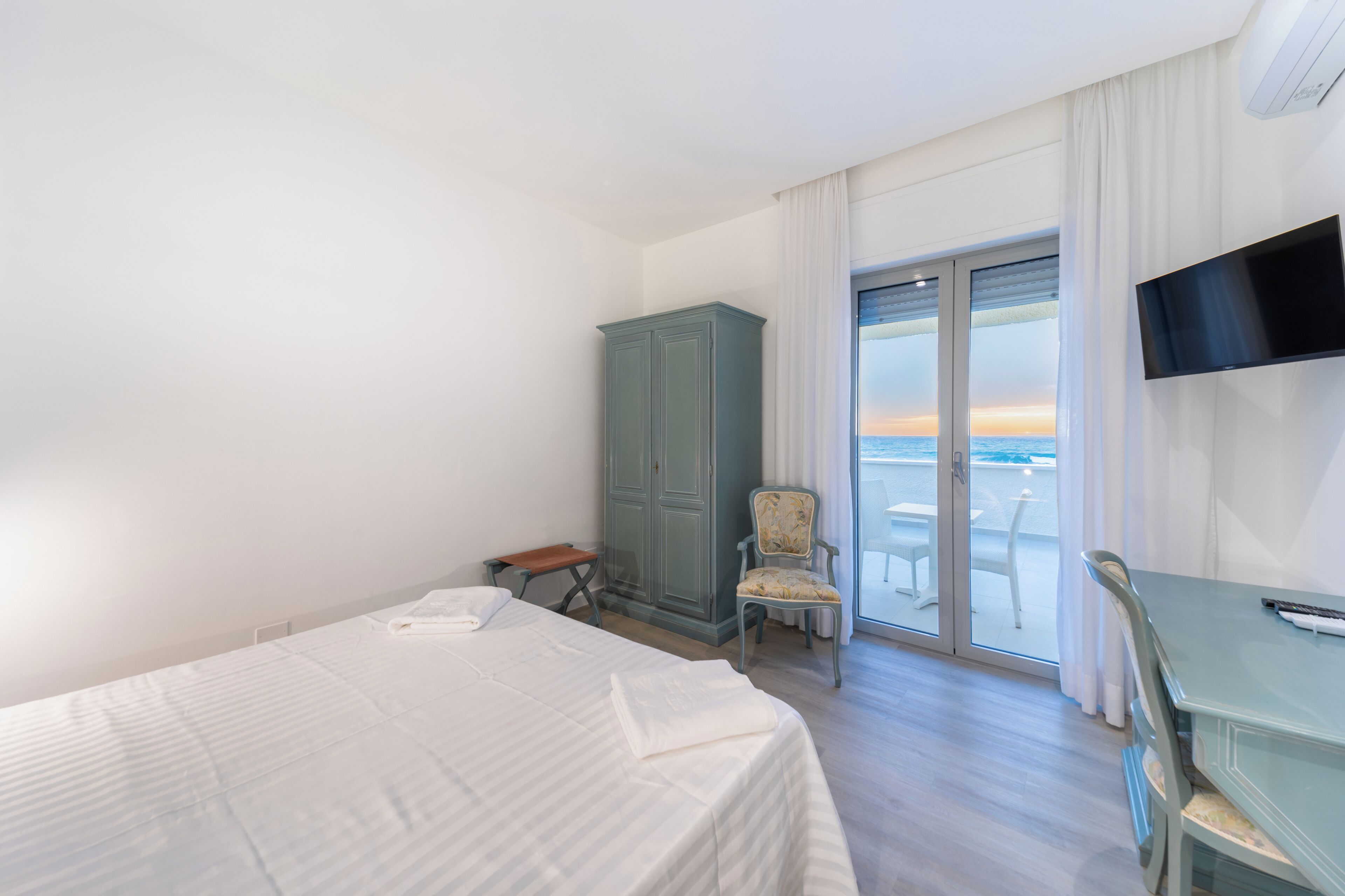 A hotel room with a bed, a chair, a TV, and a view of the sea.