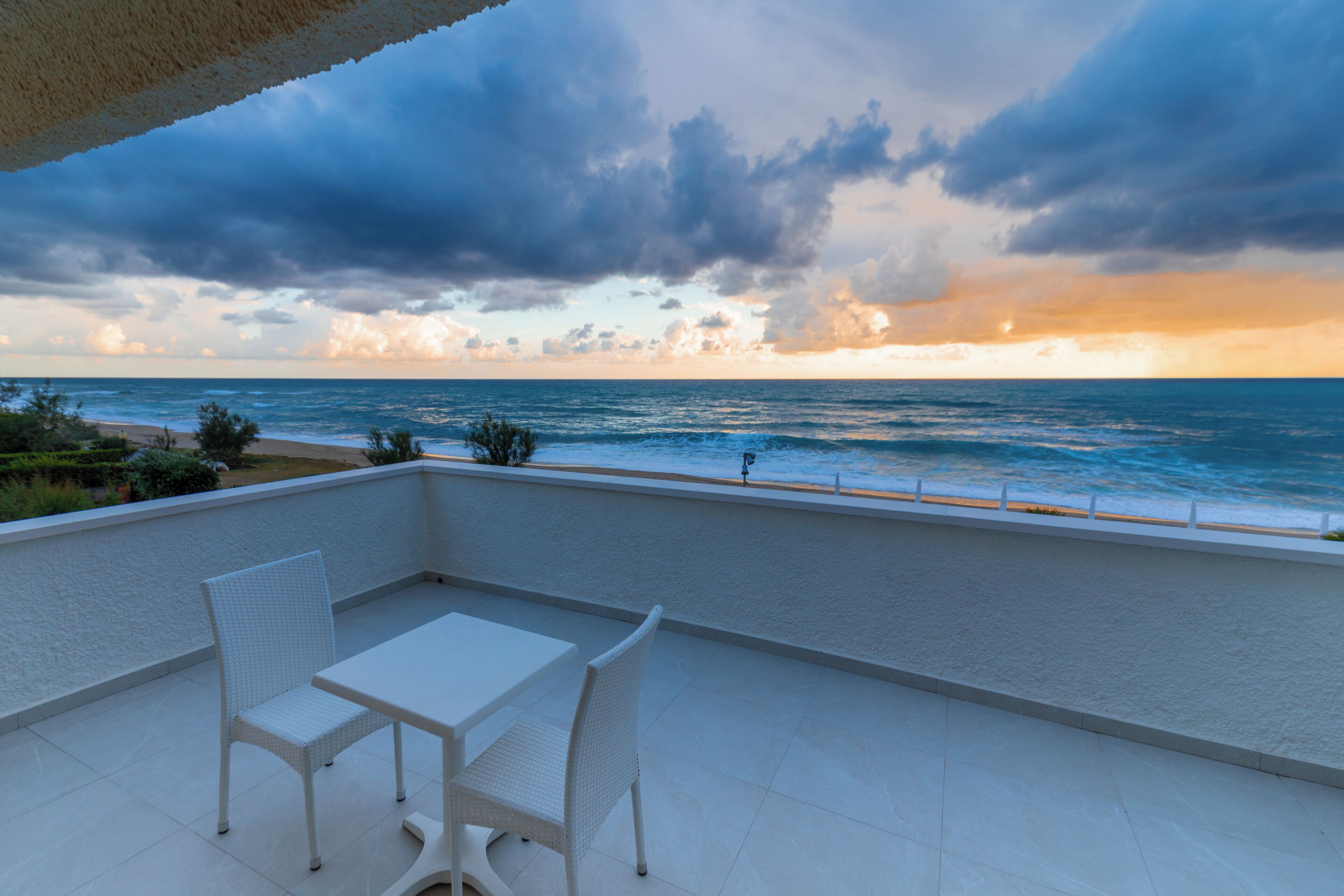 Un balcón con una mesa y sillas blancas, con vistas al mar y a la playa.